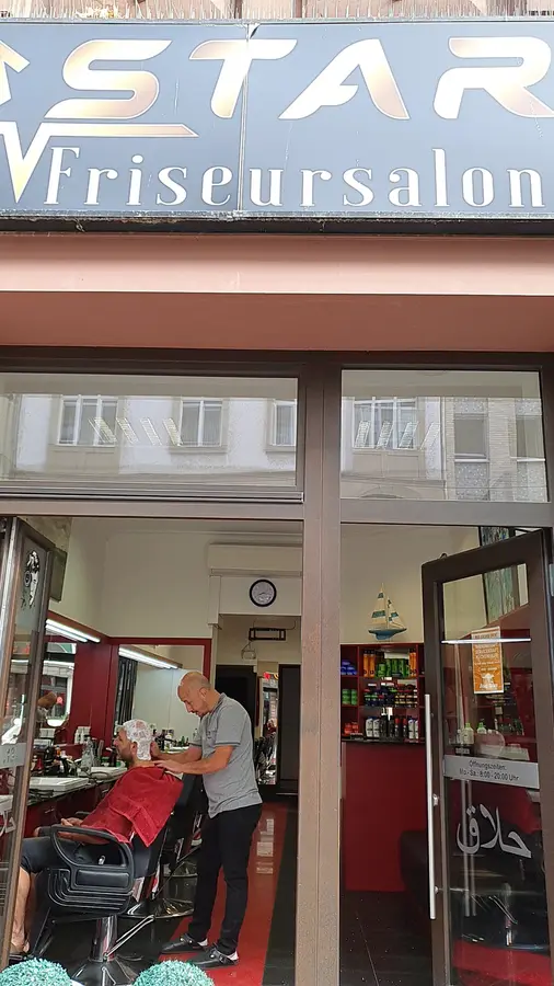 Eingang des Salons mit dem Schild STAR Friseursalon, drinnen schneidet ein Friseur einem Kunden die Haare